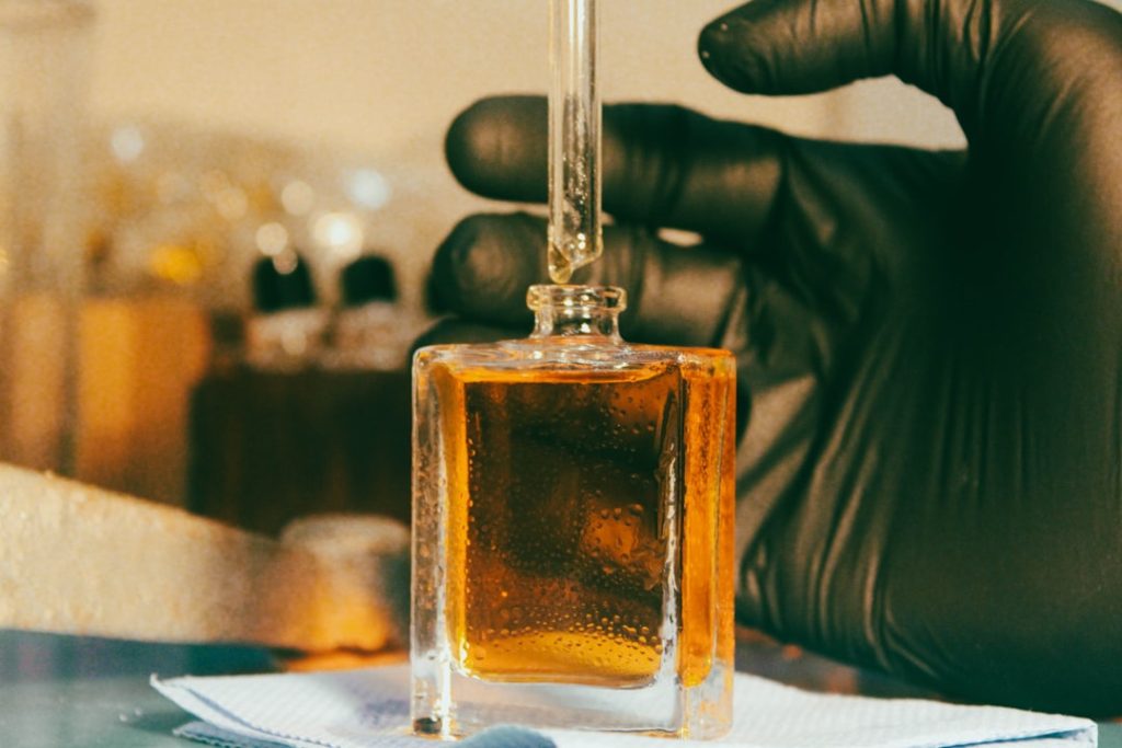 The warmth of the scene enhances the rich golden tones of the perfume within the square glass bottle. A dropper, held securely by a gloved hand, is poised to extract or dispense the scent, highlighting the artisanal process at Scentspiracy.